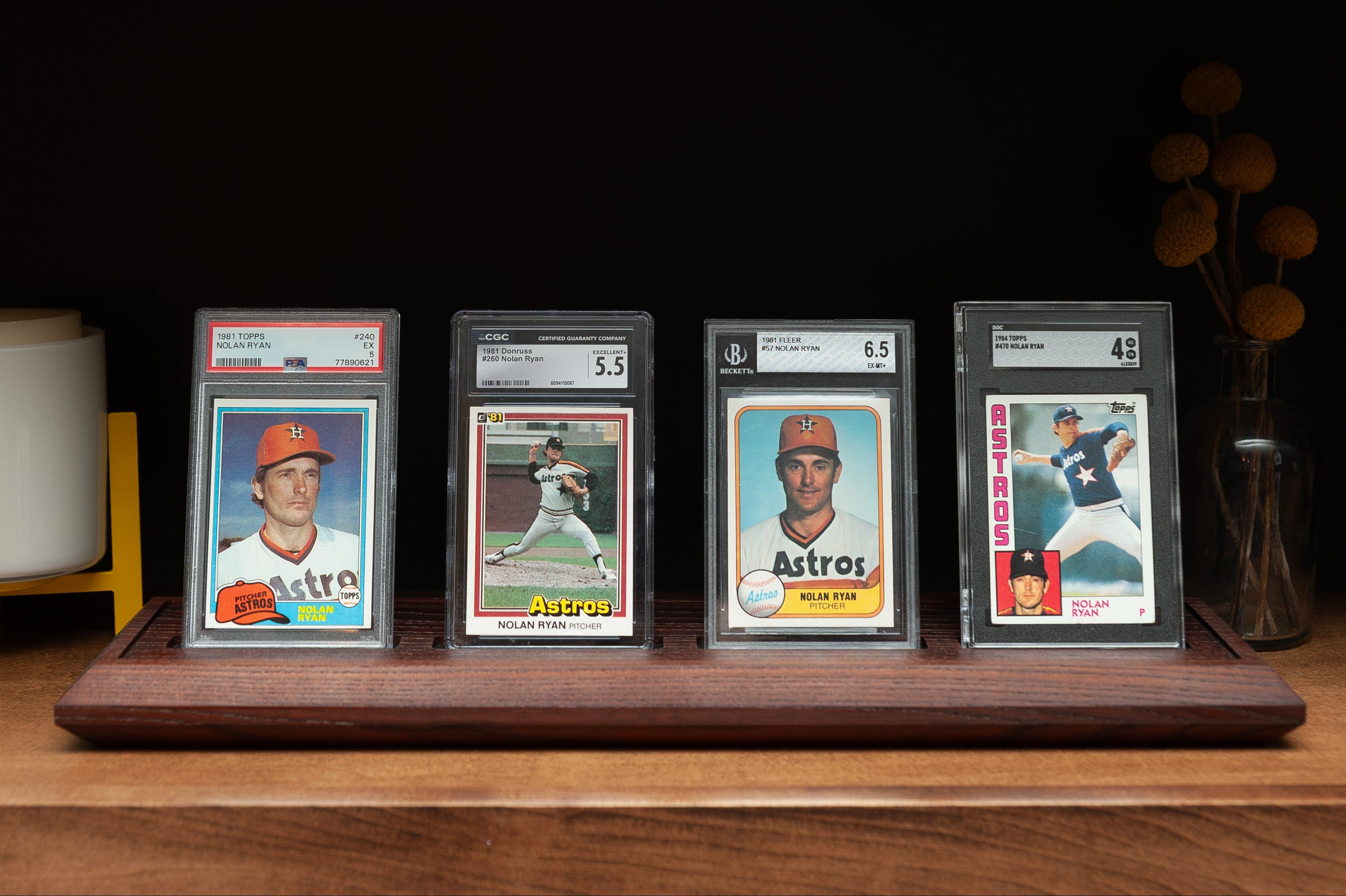 Four baseball trading cards in display cases on a wooden shelf with a dark background.